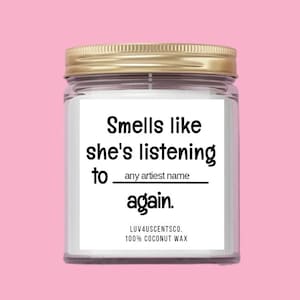May include: A white candle jar with a gold lid. The label reads "Smells like she's listening to [any artist name] again."  LUV4USCENTSCO. 100% COCONUT WAX.