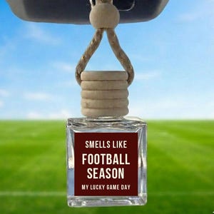 May include: Clear, square car air freshener with a brown label that reads "Smells Like Football Season, My Lucky Game Day." The freshener hangs from a beige string and wooden bead. The background is a football field.