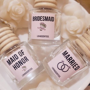 May include: Three glass car air fresheners with wooden tops. The labels are pink with black text. The labels read "Maid of Honor", "Bridemaid", and "Married".