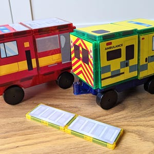 Puede incluir: Dos vehículos de juguete, un camión de bomberos rojo y una ambulancia amarilla, hechos de bloques magnéticos. Los vehículos están sobre una superficie de madera con dos bloques magnéticos adicionales.