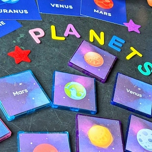Może przedstawiać: Zestaw kolorowych magnetycznych płytek z planetami i ich nazwami. Płytki znajdują się na ciemnoniebieskiej powierzchni z zestawem kolorowych liter tworzących słowo "PLANETY".
