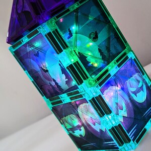 Puede incluir: Una torre de bloques de construcción magnéticos con imágenes de Halloween que brillan en la oscuridad. Los bloques son translúcidos y brillan en verde y azul. Las imágenes incluyen calabazas, murciélagos y una luna llena.
