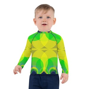 May include: A child wearing a long-sleeved shirt with a green and yellow geometric pattern. The shirt has a round neckline and a close fit.