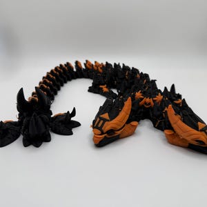 May include: A black and orange articulated dragon figurine. The dragon has a segmented body, with orange accents on the head, horns, and spine. The dragon is posed on a white surface.