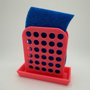 May include: A pink plastic sponge holder with a blue sponge inside. The holder has a grid pattern and a tray at the bottom.