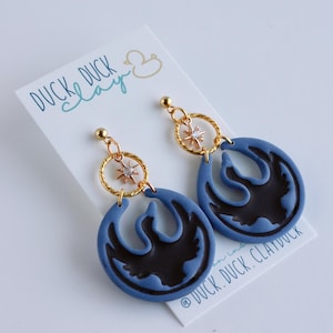 May include: A pair of blue and black drop earrings featuring a stylized bird design. The earrings have gold-colored accents, including a small star-shaped charm and a circular hoop. The earrings are displayed on a white card with the text "DUCK DUCK clay".