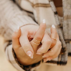 May include: A set of three thin gold rings worn on fingers. Two rings are stacked on one finger, featuring small clear stones in a delicate band design. A third matching ring with small clear stones is worn on an adjacent finger.