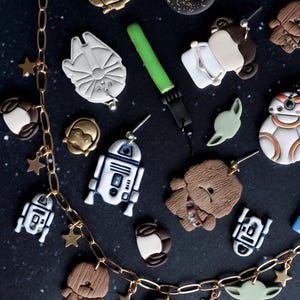 May include: A collection of handmade Star Wars-themed jewelry, including earrings and a necklace. The earrings feature characters like R2-D2, Yoda, and Chewbacca. The necklace has star charms and character pendants. The background is a dark blue with white star-like specks.