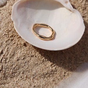 May include: A gold-toned ring with an irregular, organic, and wavy band shape. The ring features a smooth, reflective metallic surface that catches the light, creating subtle highlights and shadows along its uneven, flowing circular form.