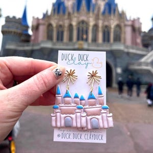 May include: A pair of pink and blue castle-shaped earrings with gold firework accents. The earrings are on a white card with the text "Duck Duck Clay" and a duck logo.