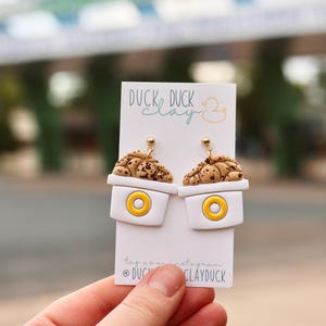 May include: A pair of dangle earrings featuring a white cup design with a yellow center circle and a textured brown speckled cookie top. The earrings are attached to a white display card with the text DUCK DUCK clay and a duck icon.