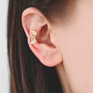 May include: A gold-colored ear cuff featuring a dragon wing design. The cuff wraps around the ear and has a small, detailed wing shape. The jewelry is worn on the ear and is a unique accessory.
