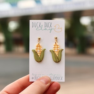 May include: A pair of dangle earrings shaped like corn cobs with yellow kernels and green husks, attached to gold-tone ball studs. The earrings are displayed on a white card featuring the text DUCK DUCK clay and a small yellow duck icon.