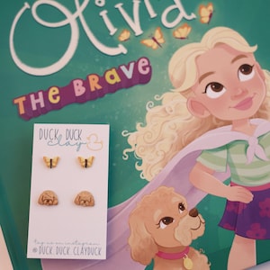 May include: A children's book cover with the title "Olivia The Brave" and illustrations of a girl and a dog. A card displays wooden earrings shaped like butterflies and dog faces. The text "Duck Duck Clay" is also visible.