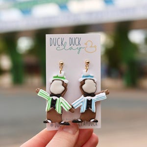 May include: A pair of dangle earrings on a white display card labeled DUCK DUCK clay. The earrings feature brown figures wearing black bow ties, black shoes, and top hats. One figure wears a green striped jacket, and the other wears a blue striped jacket.