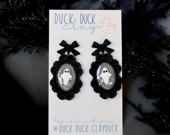 May include: A pair of black framed earrings with a ghost design. Each earring has a black bow at the top. The earrings are on a white card with the text "DUCK DUCK CLAY" and an Instagram handle.