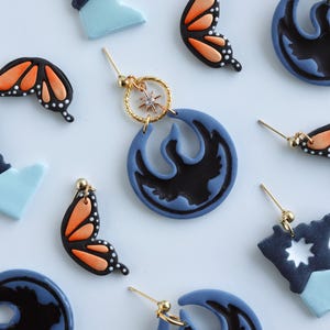 May include: Handmade polymer clay earrings in various designs. Features butterfly wings in black and orange, blue and black circular shapes, and starburst shapes. Gold-coloured earring posts and hoops.