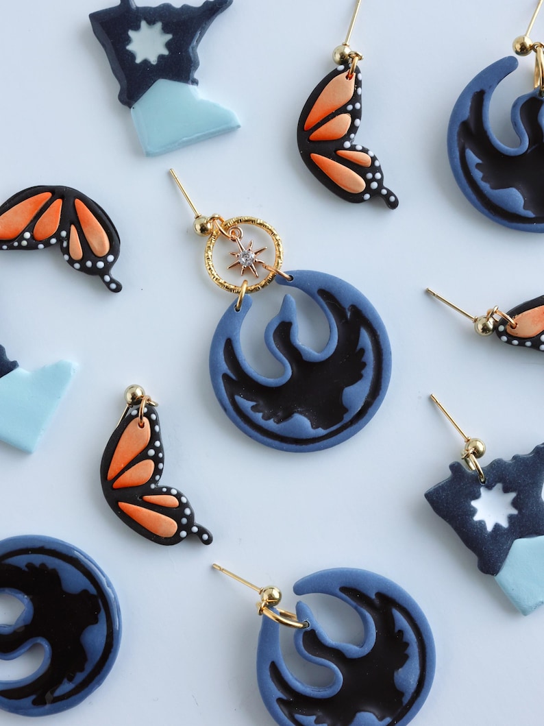 May include: Handmade polymer clay earrings in various designs. Features butterfly wings in black and orange, blue and black circular shapes, and starburst shapes. Gold-coloured earring posts and hoops.