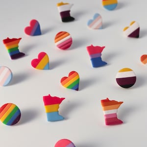 May include: A collection of small pins in various shapes including hearts, circles, and the outline of the state of Minnesota. Each pin features horizontal stripes in diverse color combinations, including rainbow, pink, yellow, blue, and purple patterns.