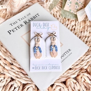 May include: Handmade bunny earrings. Each earring depicts a beige bunny wearing a blue jacket and holding a carrot. The earrings are on a white card with the text "DUCK DUCK clay" and "@DUCK.DUCK.CLAYDUCK".