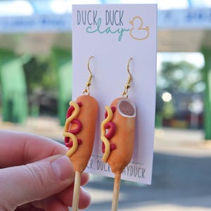 May include: A pair of dangle earrings shaped like corn dogs on a white display card with the text DUCK DUCK clay. The earrings feature orange-brown corn dog shapes with yellow mustard and red ketchup lines, attached to gold-tone metal hooks.