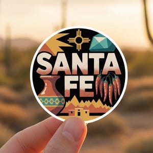 Puede incluir: Una pegatina redonda con la inscripción "SANTA FE" en blanco, con un diseño que incluye una gema turquesa, chiles rojos y motivos de cerámica nativa americana. La pegatina se sostiene frente a un paisaje desértico con cactus.