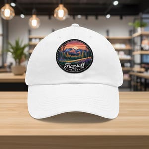 May include: White baseball cap featuring a circular patch with a landscape design. The patch includes a sunset, mountains, and trees, with the text "Flagstaff Arizona" in a curved font. The cap is made of a soft material.