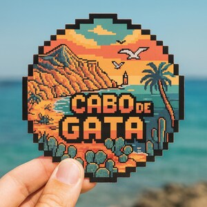 Puede incluir: Una pegatina pixelada que representa un paisaje costero con el texto "CABO DE GATA". El diseño incluye una playa, montañas, palmeras y cactus, con una paleta de colores de puesta de sol naranja, amarillo y azul. La pegatina se sostiene contra un fondo borroso del océano.