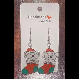 May include: A pair of dangle earrings featuring a koala wearing a Santa hat and sitting in a Christmas stocking. The earrings are attached to a white card with the text "Handmade with love".