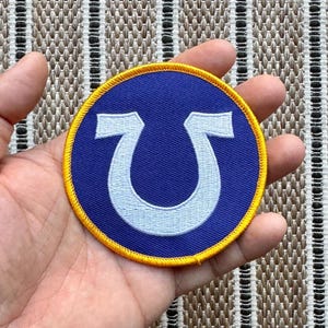 Può includere: Una toppa ricamata blu e gialla con un disegno a ferro di cavallo bianco.