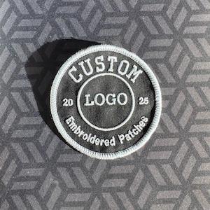 May include: A circular, black embroidered patch with white lettering and a white border. The text reads "CUSTOM LOGO" within a circle, with "20" and "25" on either side. The words "Embroidered Patches" are arched below the circle.