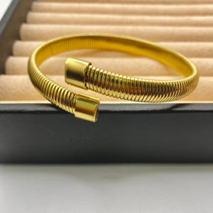May include: A gold-colored bracelet with a coiled design. The bracelet has a spring-like appearance and features a clasp closure. The jewelry is displayed on a black surface with a beige background.