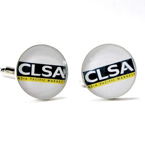 May include: A pair of round silver-toned cufflinks featuring a white background with the black text CLSA and the yellow text ASIA-PACIFIC MARKETS printed across the center. The design is encased under a smooth, clear, domed glass surface.