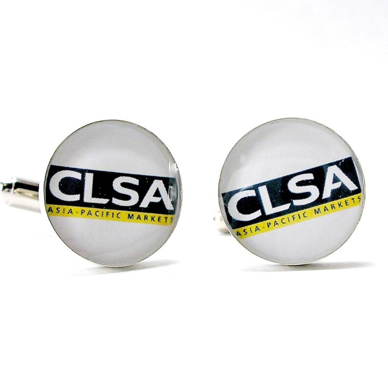 May include: A pair of round silver-toned cufflinks featuring a white background with the black text CLSA and the yellow text ASIA-PACIFIC MARKETS printed across the center. The design is encased under a smooth, clear, domed glass surface.