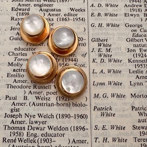 May include: Four vintage gold-tone buttons with iridescent, pearly white centers. The buttons are round with a raised edge and are set against a backdrop of aged book pages. A classic accessory.