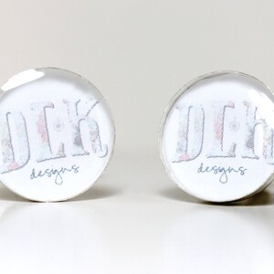 May include: A pair of round silver-toned cufflinks featuring a white background with the text "DLK" in a large, patterned font and the word "designs" in a smaller, cursive script underneath. The cufflinks have a smooth, reflective finish.