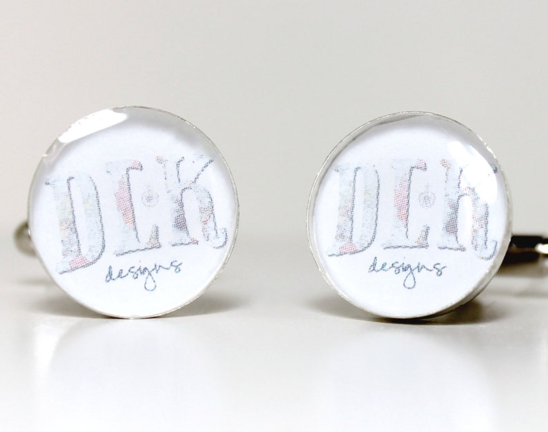 May include: A pair of round silver-toned cufflinks featuring a white background with the text "DLK" in a large, patterned font and the word "designs" in a smaller, cursive script underneath. The cufflinks have a smooth, reflective finish.