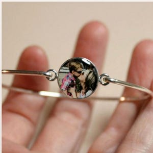 May include: A silver-toned metal bangle bracelet featuring a circular charm in the center. The charm contains a small, color photograph of two people embracing, with one person wearing a pink and white plaid shirt and sunglasses.
