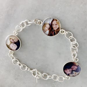 May include: A silver chain bracelet with three circular photo charms. The photos show smiling people. The bracelet has a toggle clasp and is displayed on a gray surface. The bracelet is a unique and personalized piece of jewelry.
