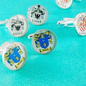 May include: A collection of silver-toned circular cufflinks featuring various colorful heraldic crests and coats of arms. Each piece displays intricate shield designs with detailed illustrations, including eagles and floral motifs, set under a clear glass dome.