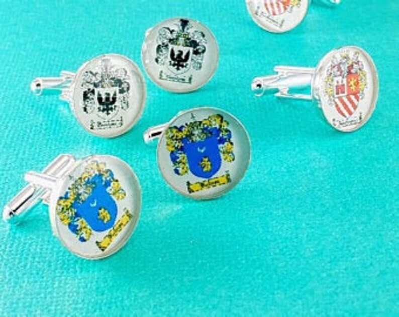 May include: A collection of silver-toned circular cufflinks featuring various colorful heraldic crests and coats of arms. Each piece displays intricate shield designs with detailed illustrations, including eagles and floral motifs, set under a clear glass dome.