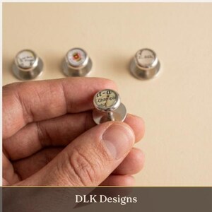May include: Four silver-toned cufflinks with circular faces featuring printed text and map-like graphics. Each cufflink has a polished metal finish and a rounded top. The text on the front-most cufflink clearly displays the word Champai.