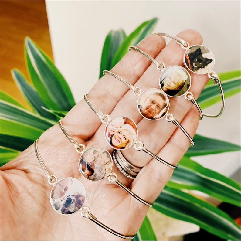 May include: Five silver-toned metal bangles, each featuring a circular charm with a custom photographic portrait. The bracelets are thin, flexible, and designed to be worn around the wrist, showcasing various images of people and a pet dog.
