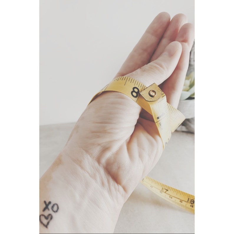 May include: A flexible yellow measuring tape wrapped around a hand, showing the number 8 clearly printed in black on the tape. The measuring tape features black increment markings and numbers, with a metal end piece visible at the measurement point.