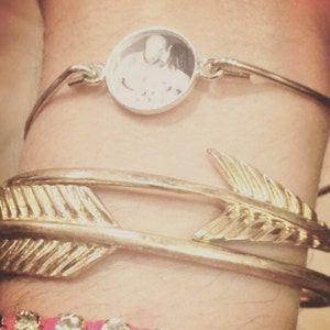 May include: A silver-toned bangle bracelet featuring a circular pendant with a black and white photograph of two people, positioned above a gold-toned bangle bracelet shaped like an arrow and a thin bracelet with small clear stones.