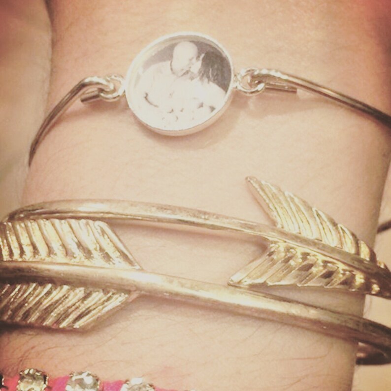 May include: A silver-toned bangle bracelet featuring a circular pendant with a black and white photograph of two people, positioned above a gold-toned bangle bracelet shaped like an arrow and a thin bracelet with small clear stones.