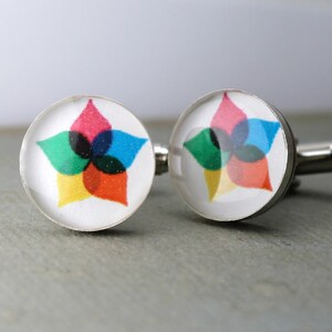 May include: A pair of round silver-toned cufflinks featuring a white background with a central graphic design. The design consists of five overlapping petal-like shapes in vibrant shades of pink, blue, green, yellow, and orange, creating a star-like pattern.
