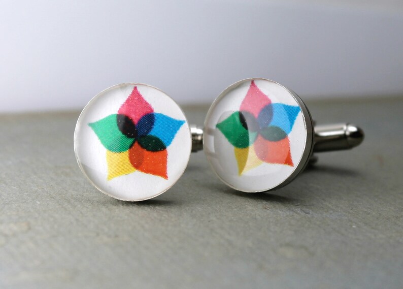 May include: A pair of round silver-toned cufflinks featuring a white background with a central graphic design. The design consists of five overlapping petal-like shapes in vibrant shades of pink, blue, green, yellow, and orange, creating a star-like pattern.