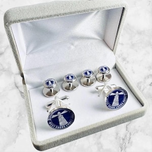 May include: A set of silver cufflinks and shirt studs in a gray velvet box. The cufflinks and studs feature a blue and white lighthouse design with the text "WEST SHORE LODGE 8064".