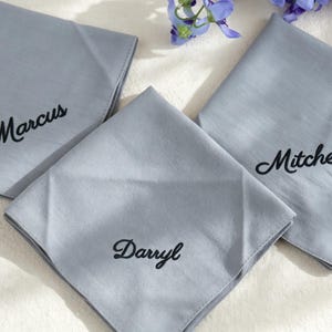 May include: Three square, light grey fabric handkerchiefs, each featuring black embroidered script text. The names Marcus, Mitchell, and Darryl are stitched onto the centre or corner of each individual cloth, which are folded neatly.